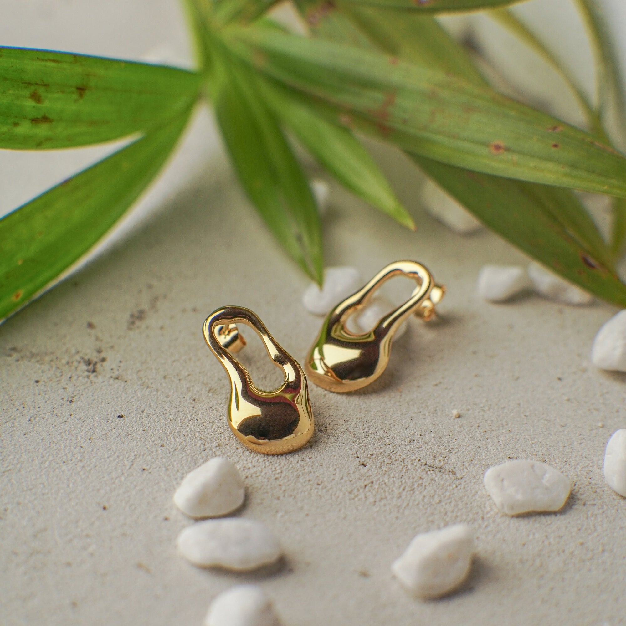 Gold earrings on a sandy surface with green leaves in the background