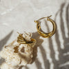 A pair of gold-colored hoop earrings placed on a textured white surface, possibly a piece of fabric or paper.
