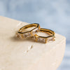 A pair of gold-colored hoop earrings with zirconia stones, displayed on a beige surface.