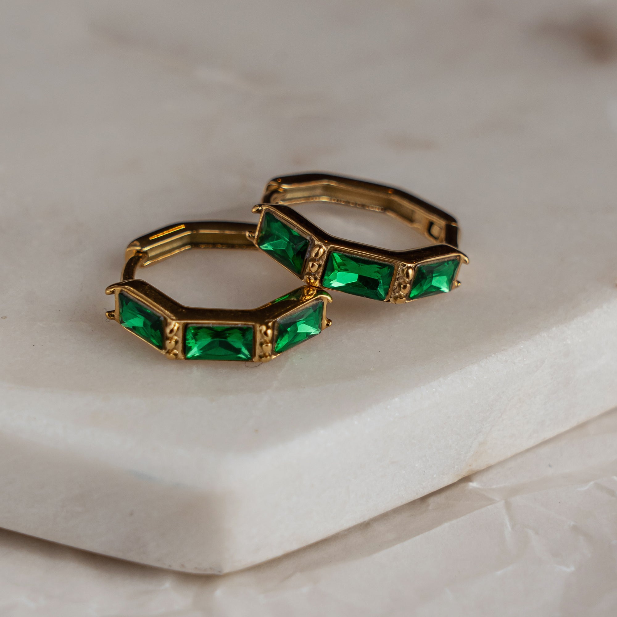 A pair of gold-colored hoop earrings with green zirconia stones set in a hexagonal pattern, placed on a white marble surface.