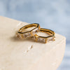 A pair of gold-colored hoop earrings with zirconia stones, displayed on a beige surface.