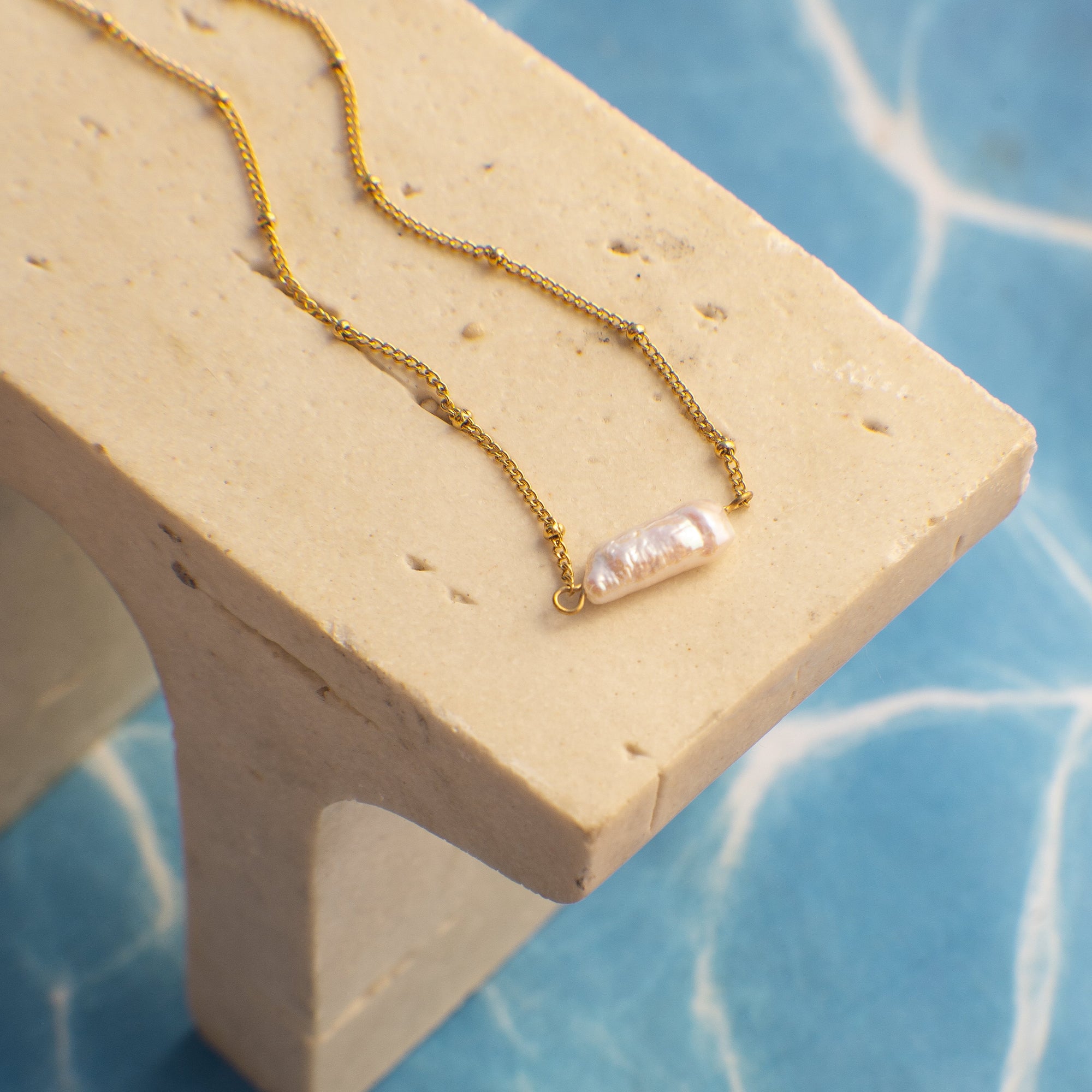 Gold necklace with a pearl pendant on a beige stand against a blue textured background