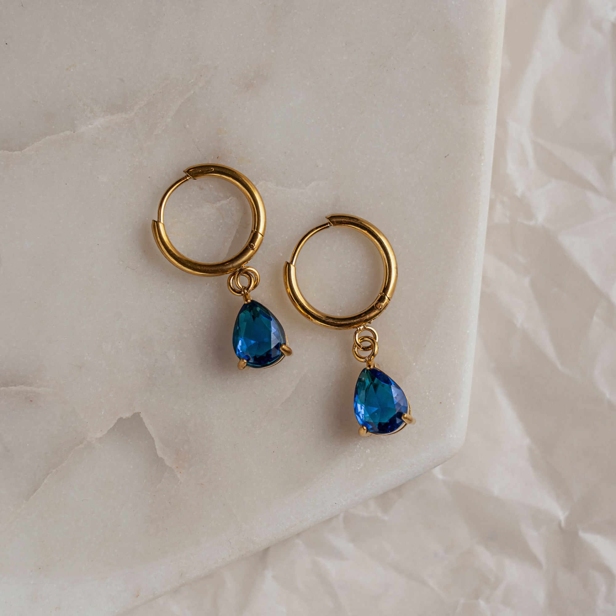 A pair of blue sapphire drop earrings with gold hoops on a marble surface.