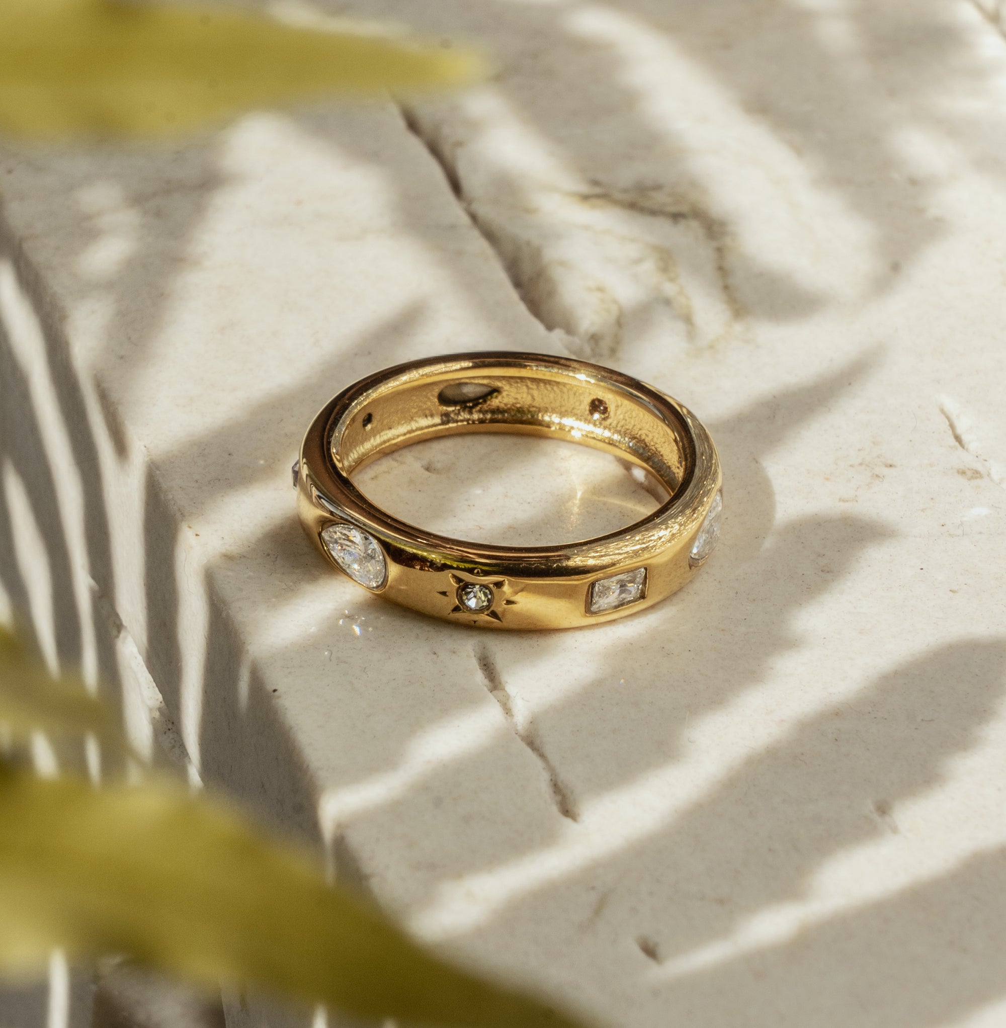 A gold ring with a zirconia center stone, placed on a textured surface with a blurred green background.