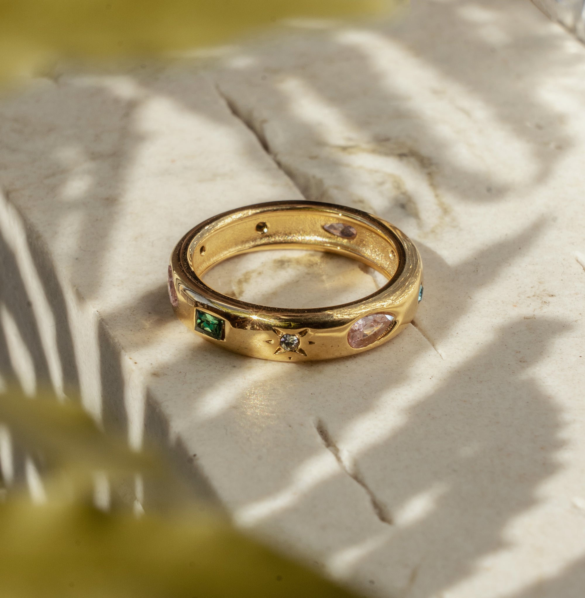 A golden ring with multicolored zirconia stones set in it, placed on a light surface with a blurred green background.