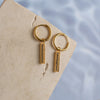 A pair of gold-colored drop earrings with zirconia pendants placed on a textured beige surface with a reflective background.