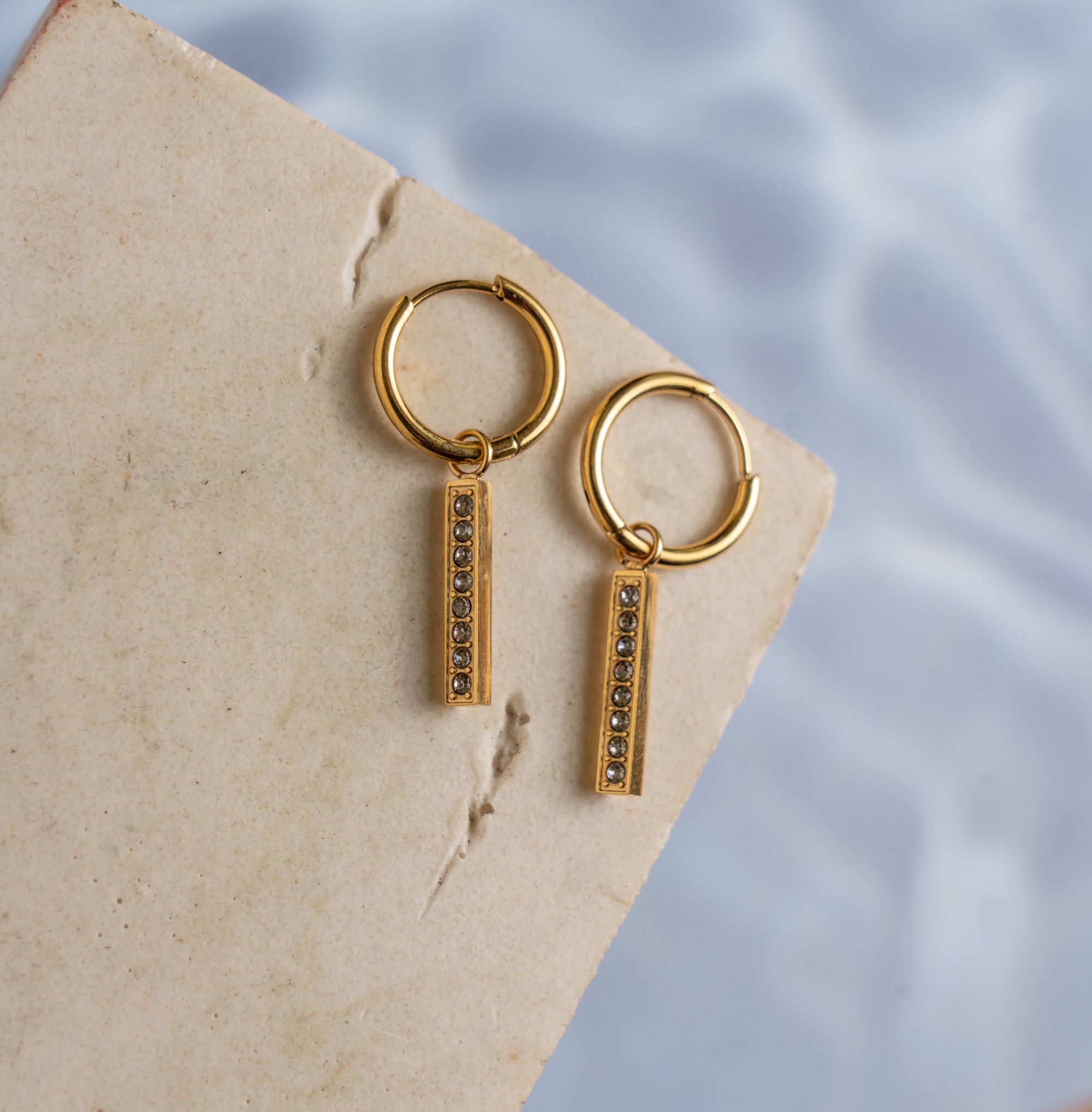 A pair of gold-colored drop earrings with zirconia pendants placed on a textured beige surface with a reflective background.