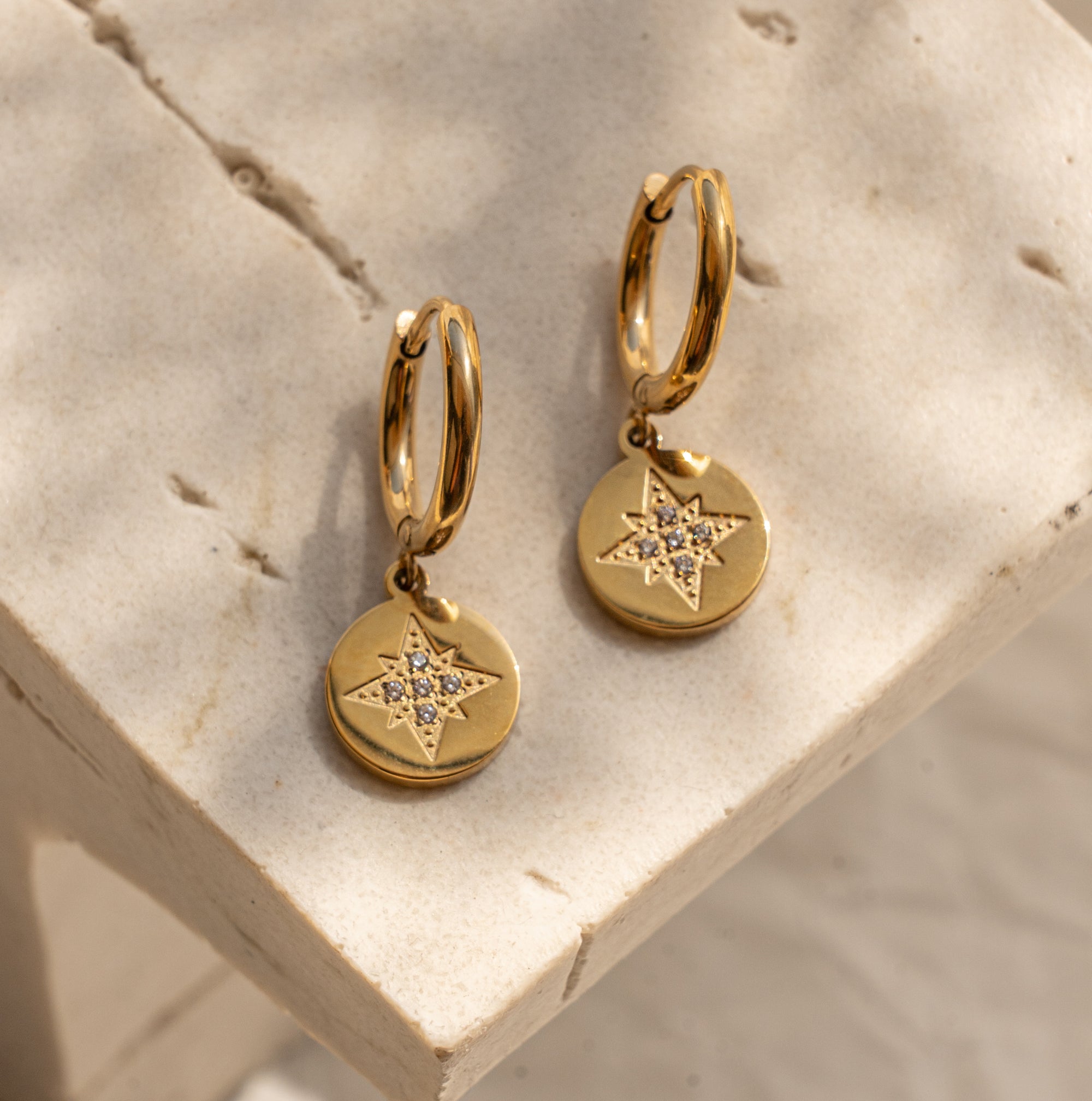 A pair of gold-colored Huggie earrings with a star drop design featuring cubic zirconia embellishments, displayed on a stone surface.