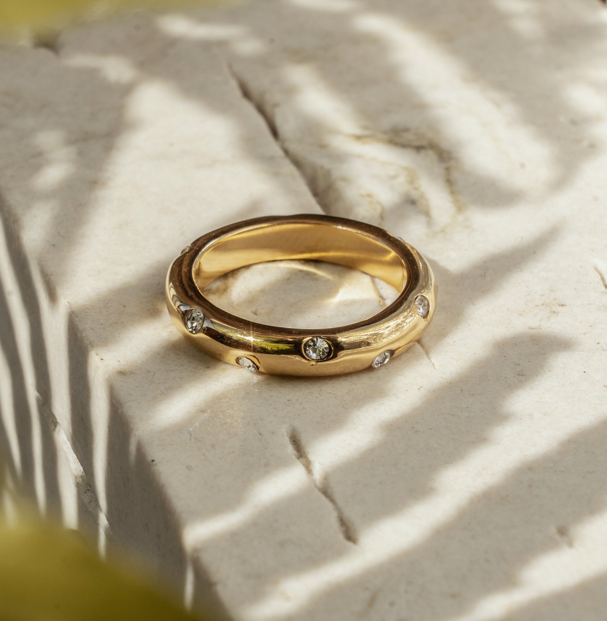 A gold ring with clear zirconia stones set around its circumference, placed on a textured surface with a blurred background.