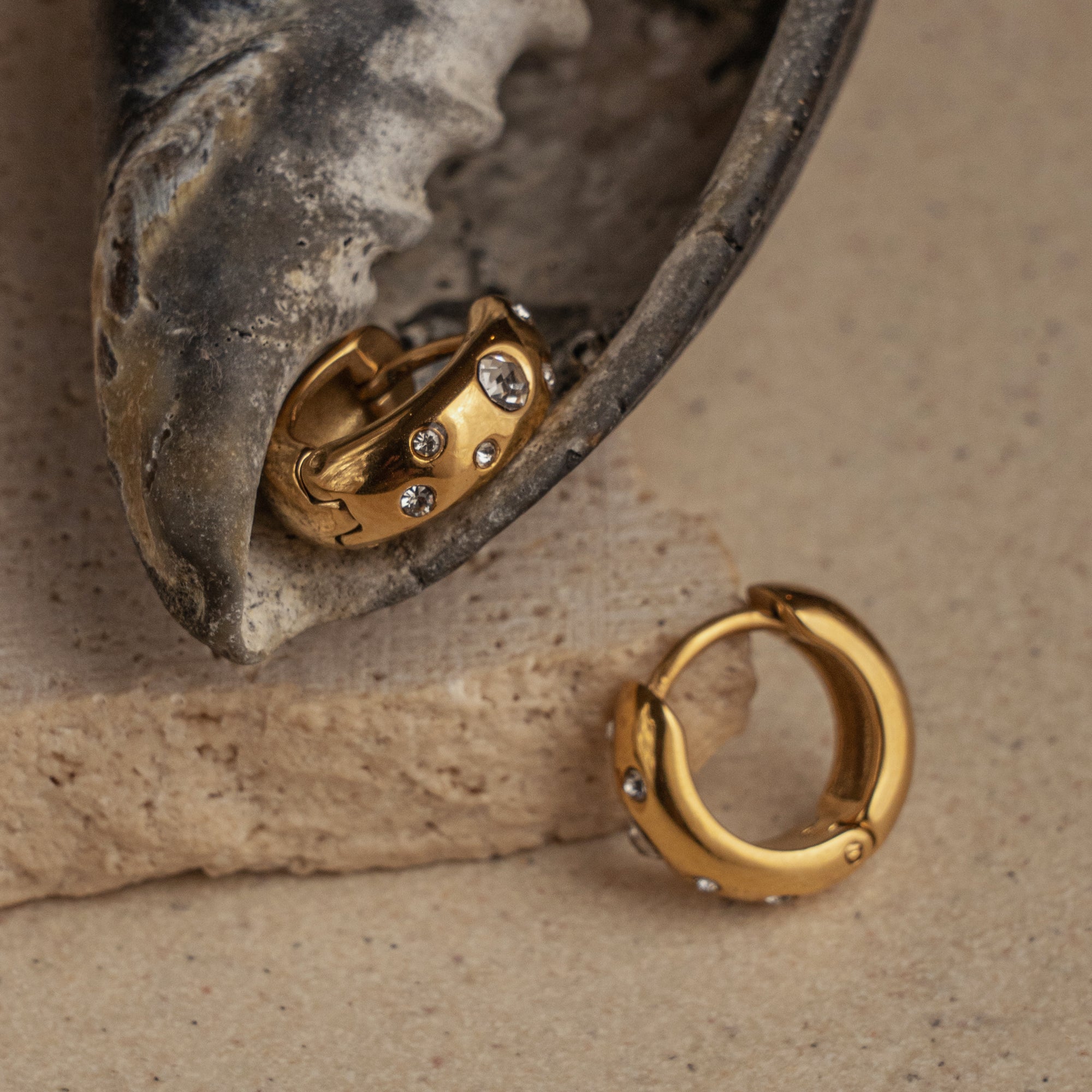 A pair of gold huggie earrings with small cubic zirconia embellishments, displayed on a textured surface.