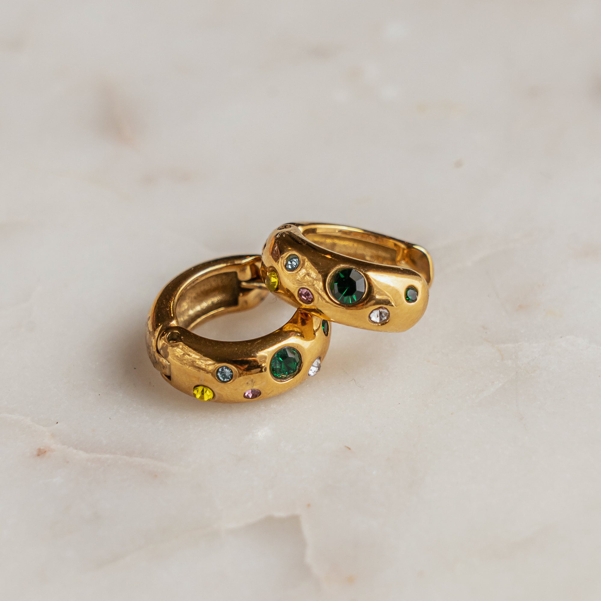 A pair of gold-colored multi-colored zirconia huggie earrings on a marble surface.