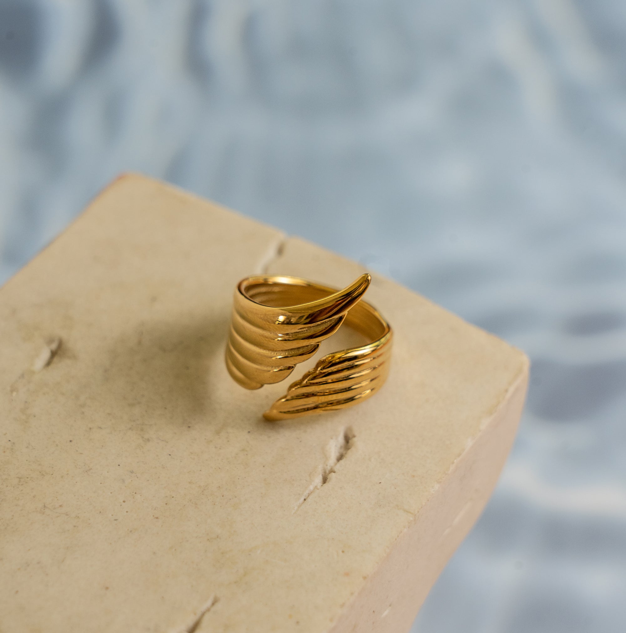 A gold-colored ring with a unique wing design, displayed on a beige surface with water drops in the background.