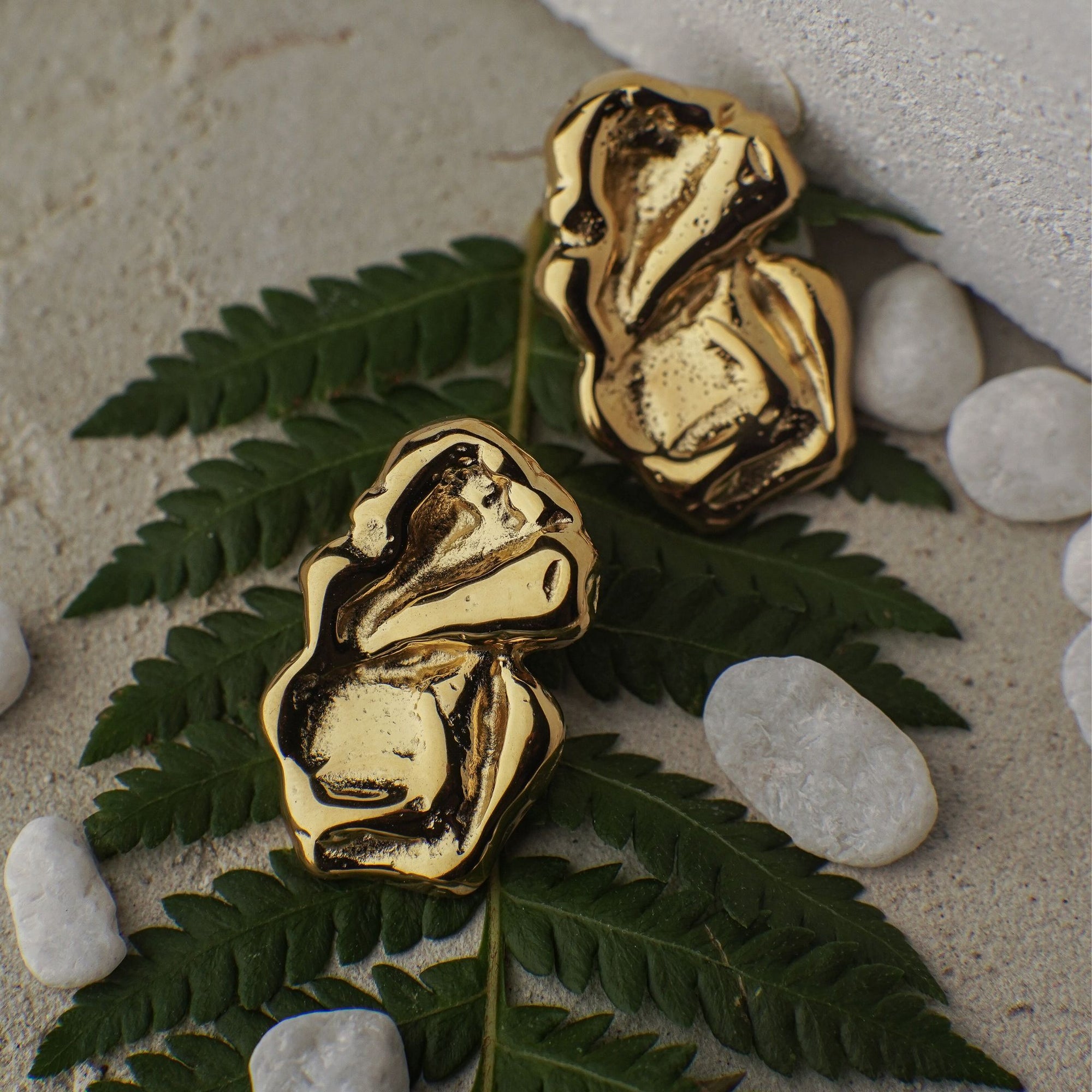 Gold rock-shaped earrings on green leaves with white stones in the background
