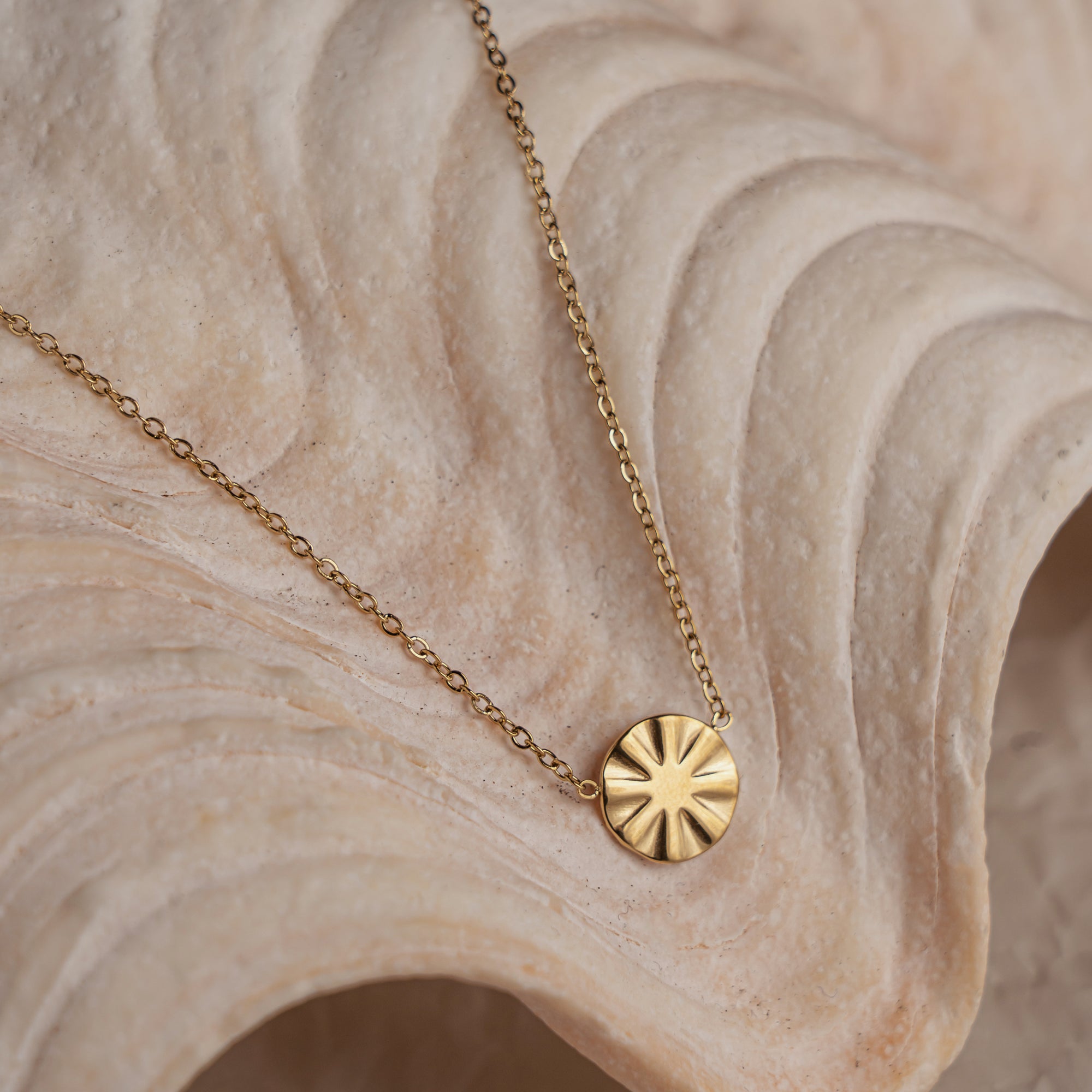A gold-colored necklace with a round pendant featuring a geometric design, displayed on a textured background.