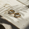 A pair of gold-colored hoop earrings with zirconia stones embedded on the hoop, placed on a marble surface.