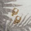 A pair of gold-colored drop earrings laid on a crumpled silver surface with a shadow of a plant leaf on them.