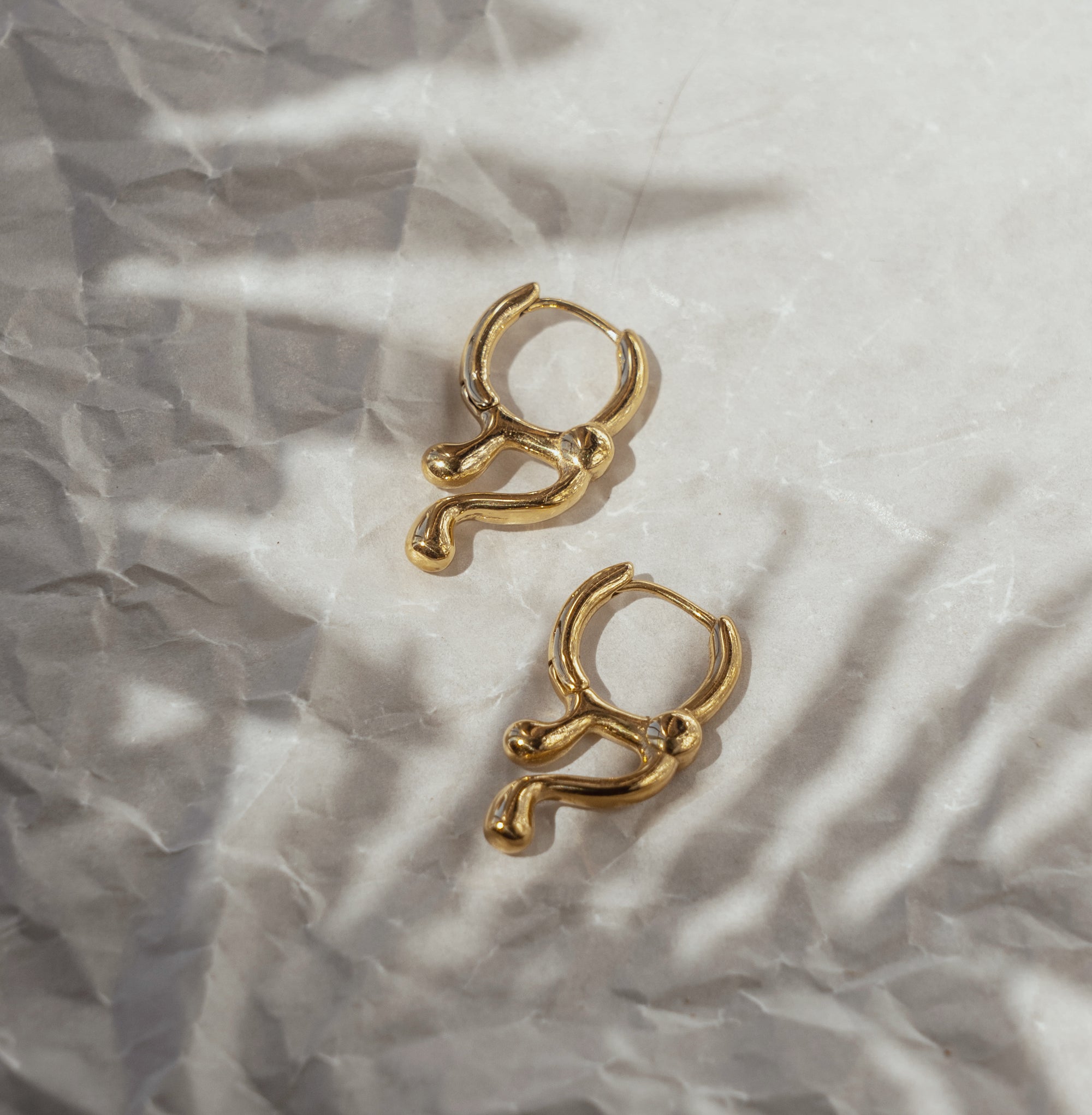 A pair of gold-colored drop earrings laid on a crumpled silver surface with a shadow of a plant leaf on them.