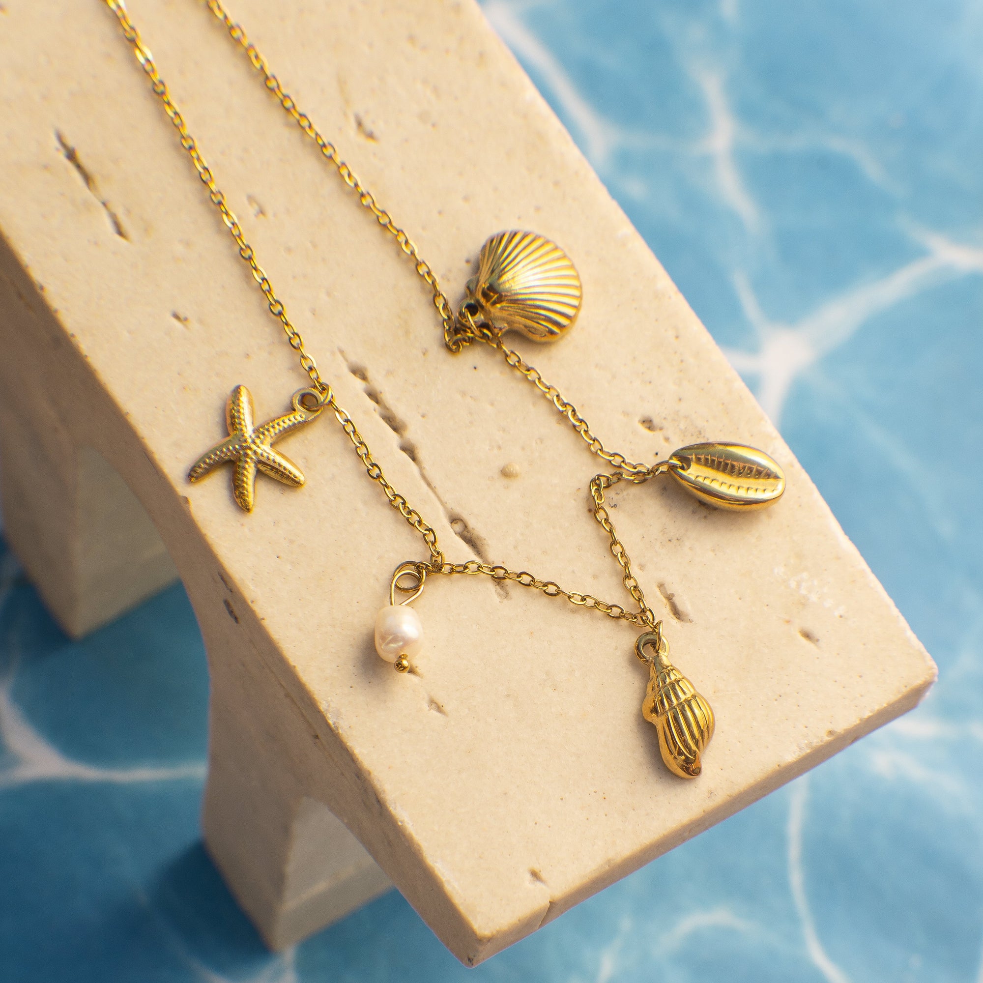 Gold necklace with starfish, shell, and hand-shaped charms on a beige stand against a blue water background
