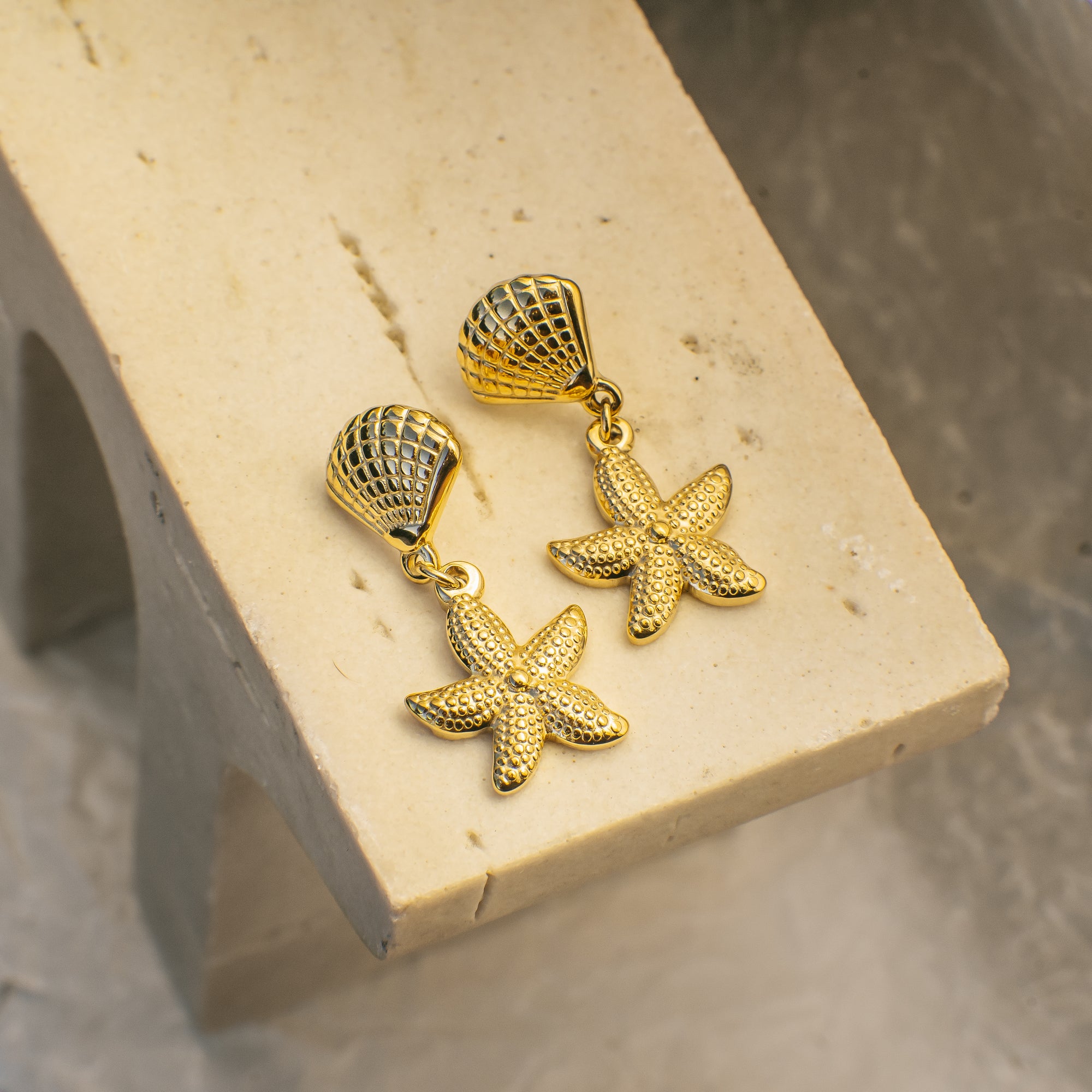 Gold starfish and shell earrings on a beige stand with a neutral background
